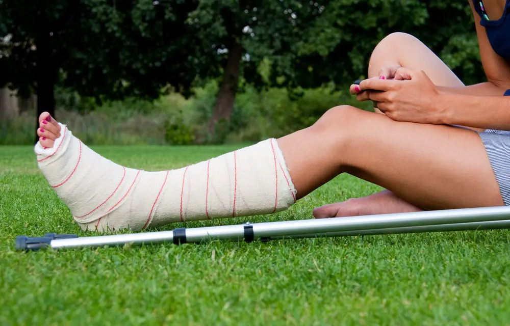 Woman sitting in grass with a cast on her leg and a crutch beside her