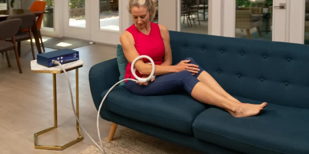 Women sits on couch and applies butterfly coil onto her elbow from TeslaFit Plus 2 PEMF Machine
